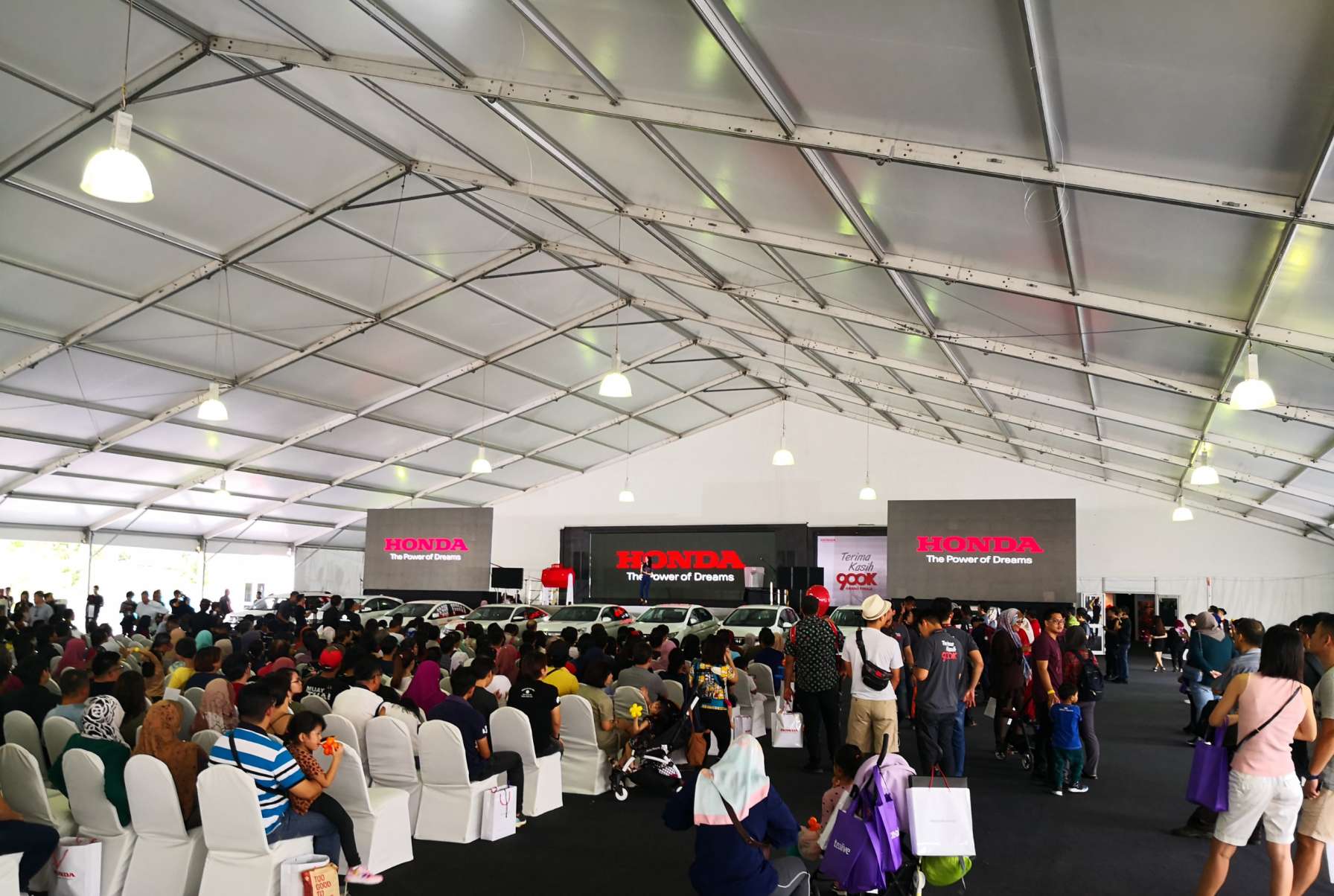 The community watches the Honda cars being showcased in front of the LED screen.