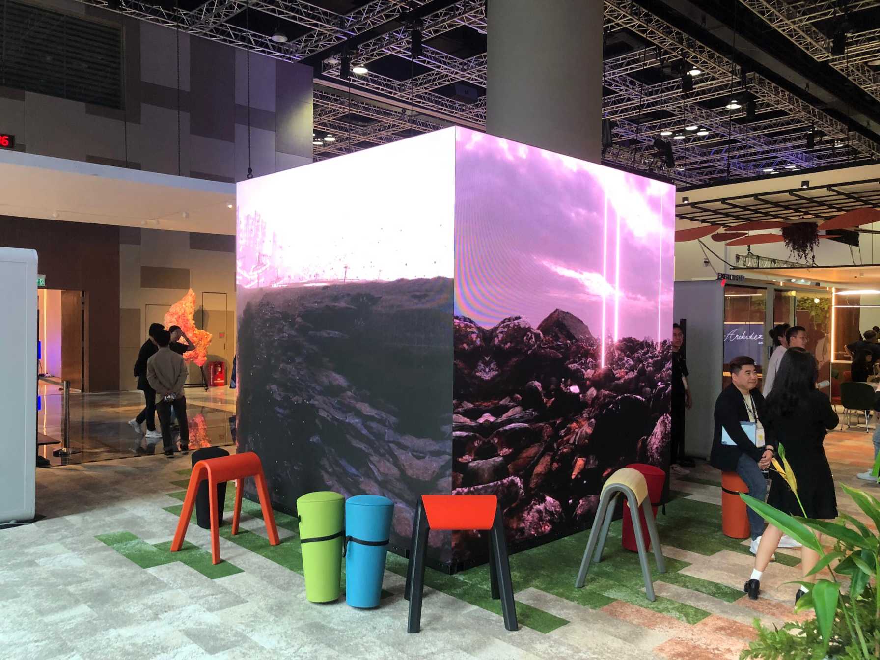 A large 3D LED cube video wall installed inside a high-ceiling exhibition hall. The seamless, multi-sided LED screens display a vibrant purple-and-pink landscape graphic, surrounded by modern seating and event attendees.