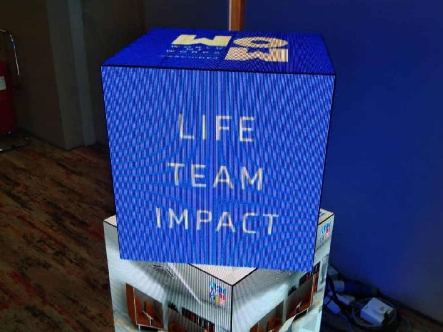 A multi-sided cubic LED digital display mounted on a base. The blue screen displays the white text "LIFE TEAM IMPACT" on the front panel and graphic logos on the top face.